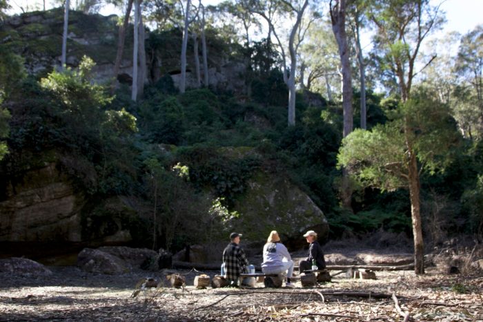 Three people sitting in a forest clearing, talking to each other.