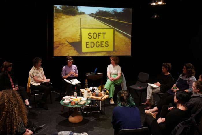 People sitting in a circle, listening to a woman talking. There is an image projected behind the woman.