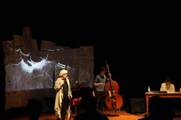 A woman on the left is speaking into a microphone, a man in the middle is playing the cello, and another man on the right is sitting at a desk with a soundboard and laptop.