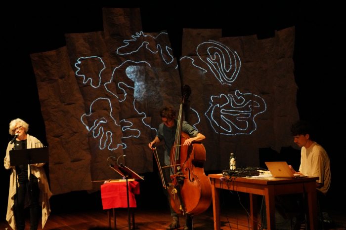 A woman on the left is speaking into a microphone, a man in the middle is playing the cello, and another man on the right is sitting at a desk with a soundboard and laptop.