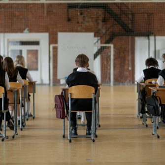 Three rows of teenagers in school uniforms are seated at wooden school desks in a large hall. They are facing away from the camera so we can only see their backs. 