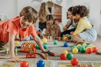 A boy in orange shirt plays on floor at front of frame while an adult instructs 3 children at the back of frame