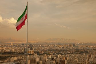 Iran flag waving  in the wind above skyline of Tehran lit by orange glow of sunset.