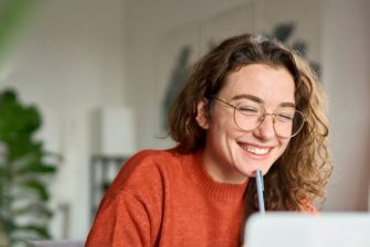 Happy young woman using laptop sitting at desk writing notes while watching webinar, studying online, looking at pc screen learning web classes or having virtual call meeting remote working from home.