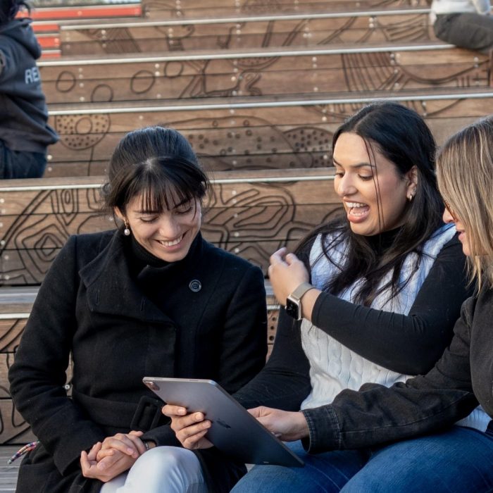 Three female students sitting on the outdoor auditorium looking at a iPad