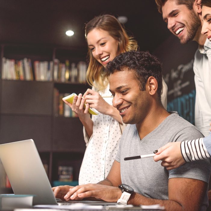 Laughing and learning. Cropped image of African student writing something on laptop while sitting at desk with his friends behind him