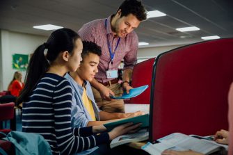 Students are working on computers with textbooks. A male teacher is standing with them, helping them with their work.