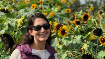 A woman smiles at the camera with a field of sunflowers behind her