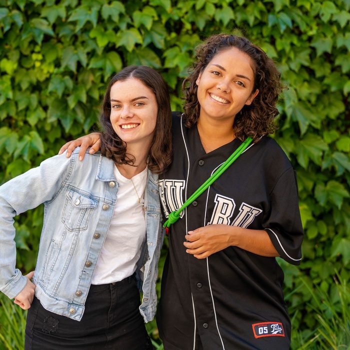 Two students with their arms around each other standing in front of a wall covered in foliage