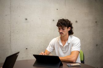 Students using their laptops at an indoor table on campus with a large blank concrete wall behind them