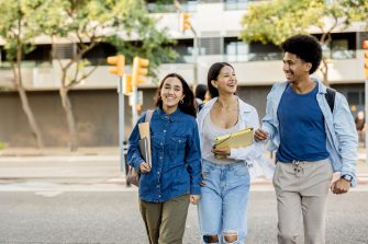 Three diverse college friends walking arm in arm, laughing and carrying books and backpacks on a sunny day.