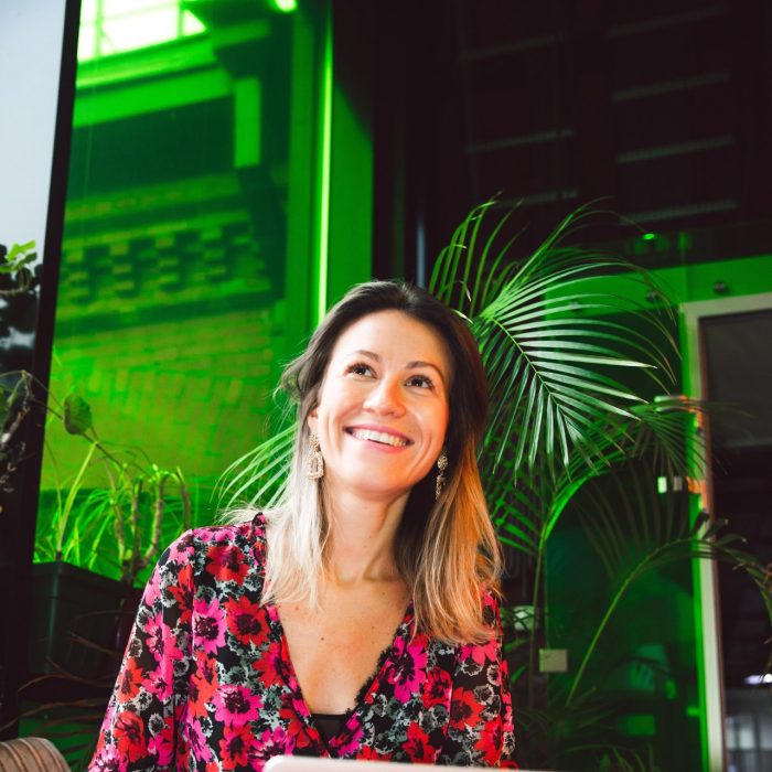 A woman sits with a laptop on her knee, smiling off camera