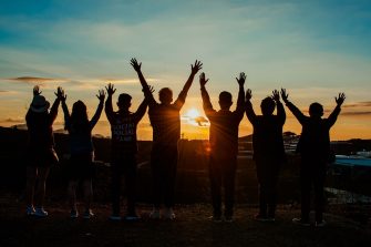 Seven people are standing in a line. They are facing the sunset with both arms up in the air.