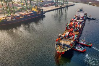 This image depicts two large shipping vessels just off port in the ocean. The image is taken from a aerial view. Each vessel has shipping containers loaded on to it.