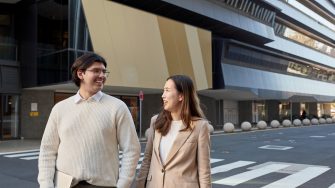 Female in pink jacket and male in beige sweater, law students, walking together behind the law building in bright, even lighting. 