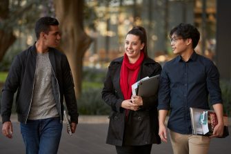 Three law students walking in front of Law Building, walking towards the camera, greenary and reflections in glass of Law Building in background