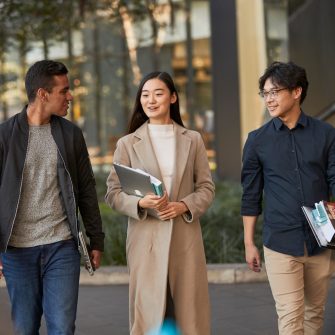 Three law students walking in front of Law Building, walking towards the camera, greenary and reflections in glass of Law Building in background