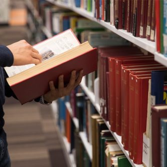 Close up of a person's hand holding an open book, they are in front of a row of book shelves.