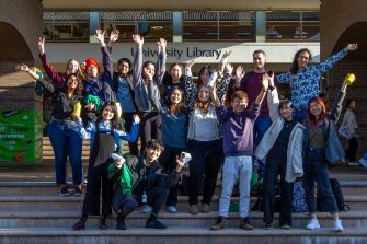 Library Peer Mentors group standing at front of Main Library (two rows plus one crouching), smiling and hands in the air with University Library signage in background.