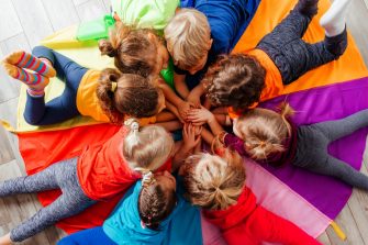 Lovely children laying on multicolor canopy in circle. Team building game for daycare children in colorful t-shirts. Top view kids on wooden floor.