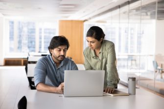 Two Indian business partners meeting at workplace table, talking at laptop, looking at screen, speaking, discussing online startup content, Internet marketing strategy in co-working office