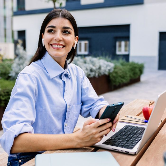 Happy entrepreneur woman with laptop using cell phone sitting outside of office building. Business lifestyle concept.