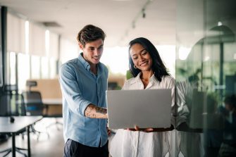 Two happy diverse multiethnic business team people working, talking in corporate office using laptop computer. Busy business man and woman colleagues at workplace.