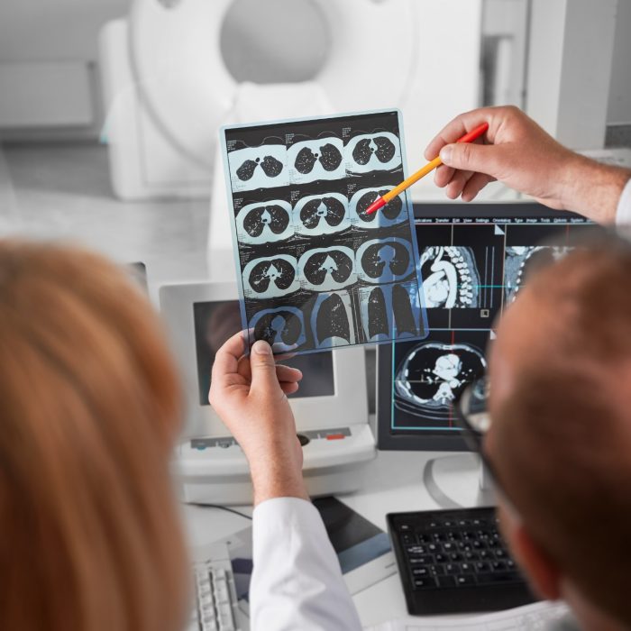 Medical computed tomography or MRI scanner. Back view of doctor and nurse sitting at computer, looking at MRI results. Doctor holding scan, showing, pointing by pen, studying, examining.