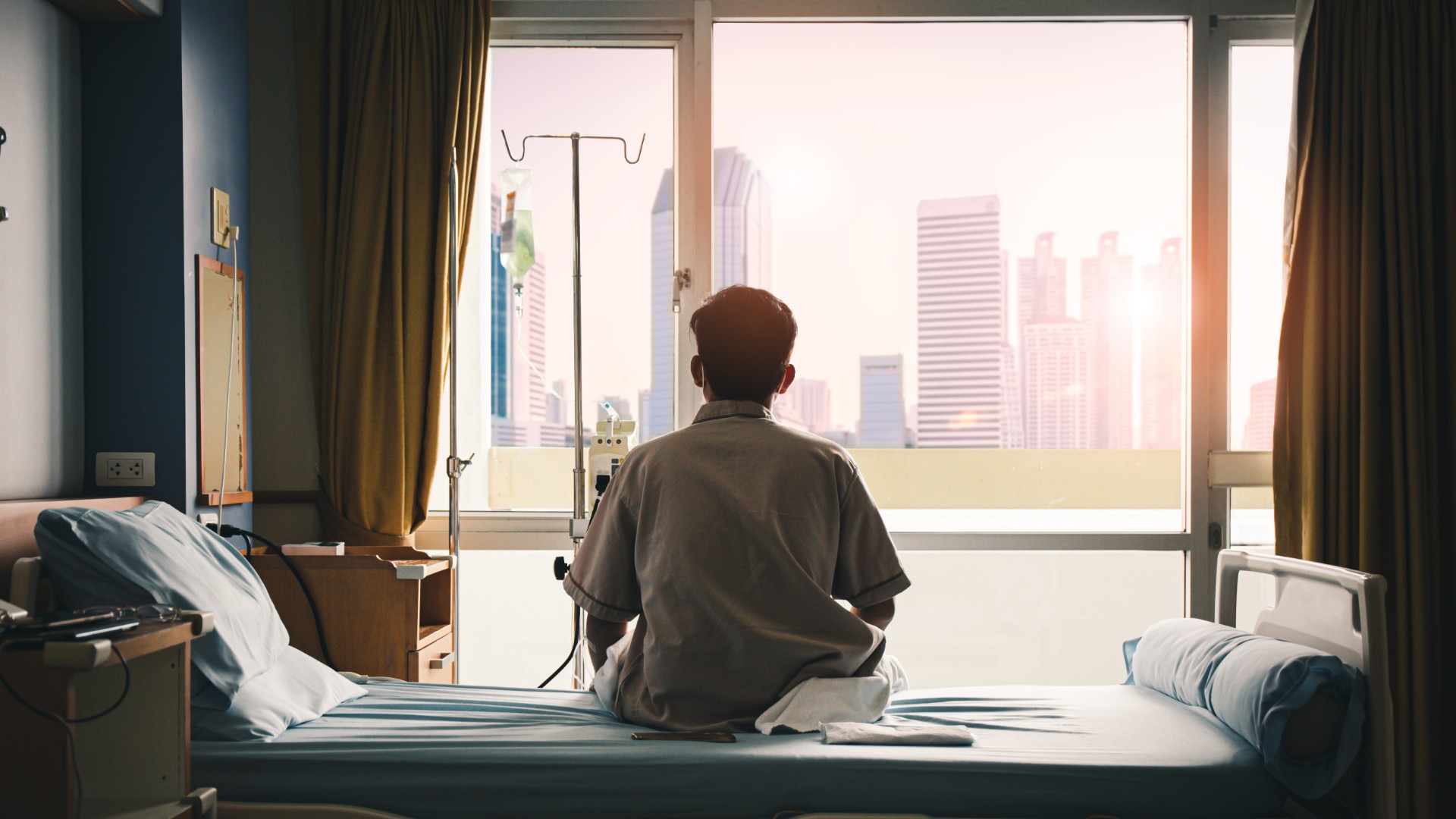A man sits on a hospital bed, looking out the window. 