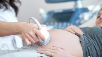 Close up of incognito professional doctor using ultrasound equipment, screening of pregnant woman. Doctor in white uniform touching stomach by tool of young female, who lying on bed at cabinet.