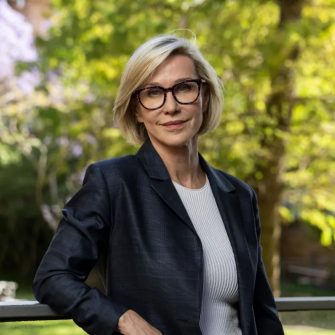 Portrait of a blonde woman with black framed glasses wearing a dark blazer with a light grey cable knit shirt underneath