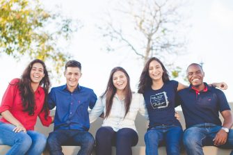 Five happy people sitting on a bench with trees and a blue sky