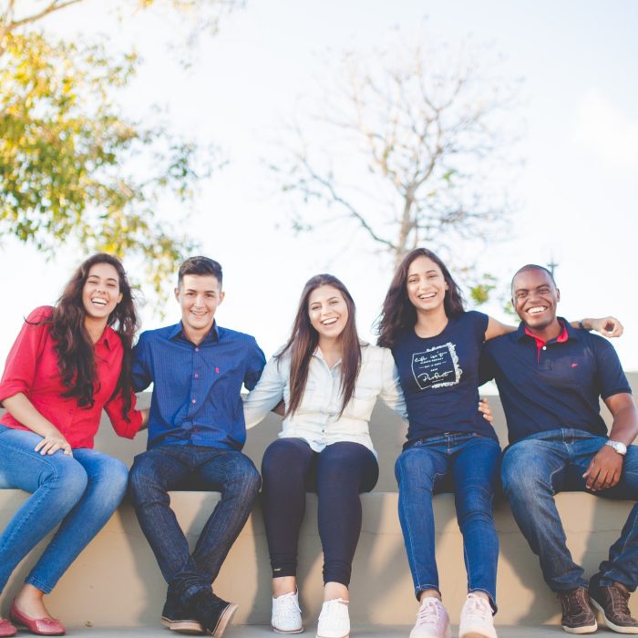 Five happy people sitting on a bench with trees and a blue sky