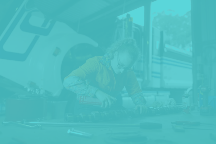 Person in a high-visibility orange work shirt using a tool on a heavy vehicle part at a workshop bench, with large truck components in the background.