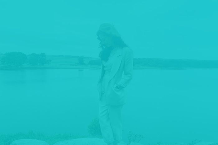 Stylized image of a person standing outdoors near a calm body of water with trees and open land in the background. The person is wearing a light-colored suit and a beret, with hands in pockets, and the entire image is overlaid with a teal color filter.