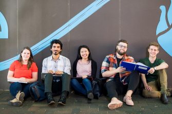 Five students sitting on the ground against a wall smiling