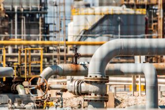 Close-up of industrial pipes and valve system inside refinery with metal structures and processing equipment in background. 