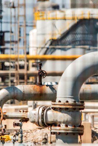 Close-up of industrial pipes and valve system inside refinery with metal structures and processing equipment in background. 