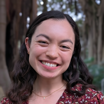 Headshot Image of Zoe-Mei Duffin-Pruksapun smiling in a paisley printed dark red dress standing in front of trees that are blurred