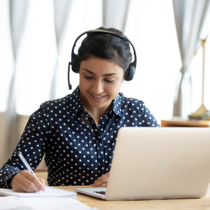 Happy indian girl student wear headset study online with webcam teacher write notes, happy young woman listen lecture watch webinar on laptop sit at desk, distance e learn language education concept