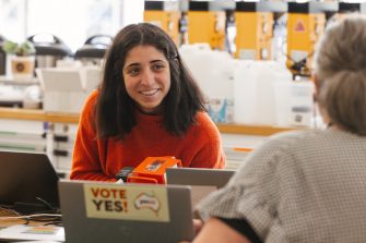 Dark haired woman in orange jumper smiling at another person who is facing her
