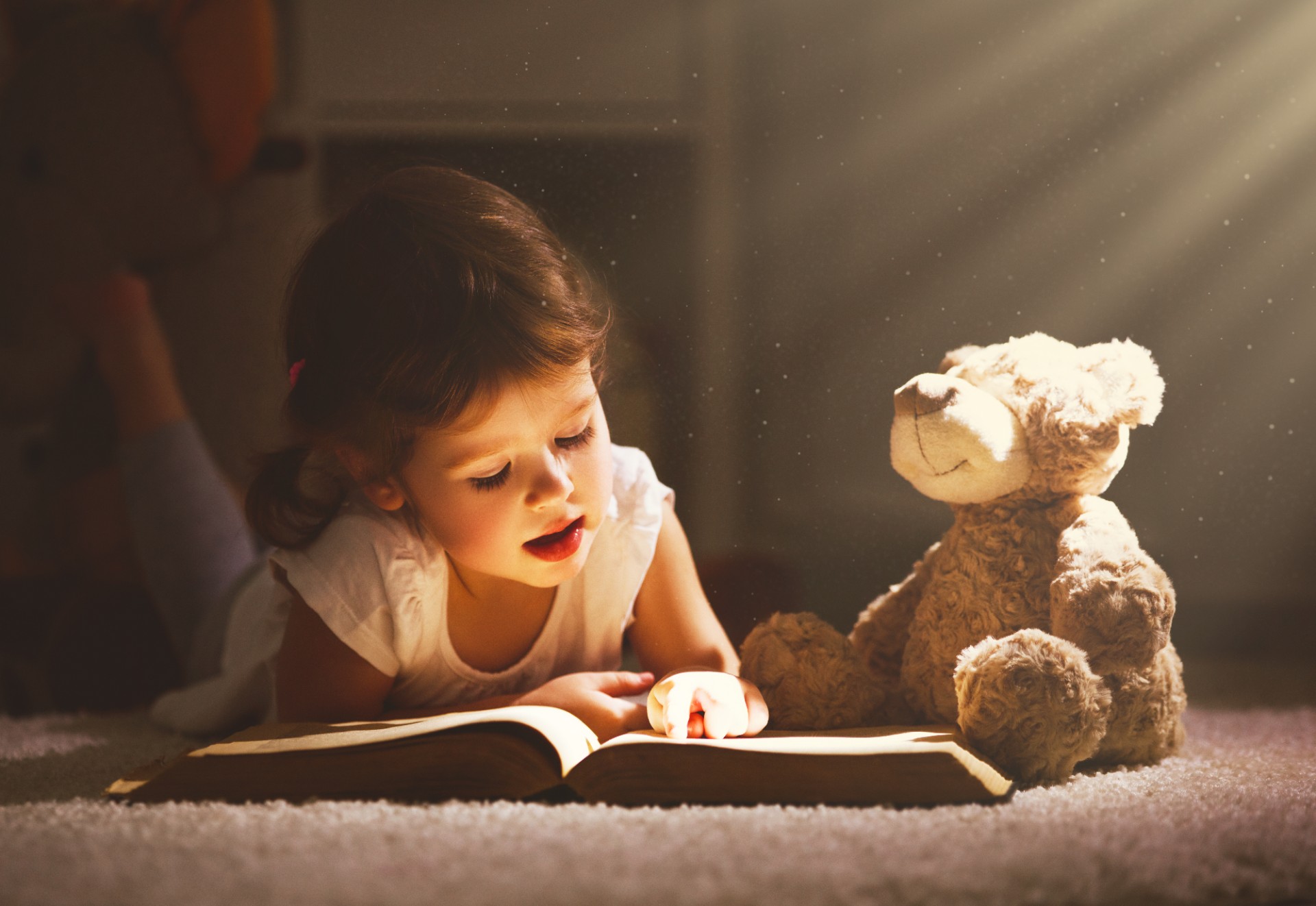 A child reading a book in evening in dark with a toy bear
