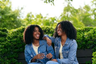 two happy twin sisters are sitting on a park bench, wearing denim jackets and laughing together on a sunny day