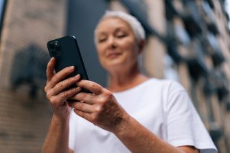 Selective focus of gray-haired senior lady using smartphone texting message standing on urban street on summertime. Middle aged woman grandma hold phone typing sms enjoying communication in mobile app