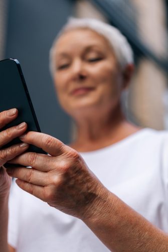 Selective focus of gray-haired senior lady using smartphone texting message standing on urban street on summertime. Middle aged woman grandma hold phone typing sms enjoying communication in mobile app