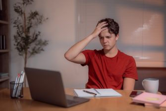 Young man touching his head with his hand while looking at his laptop, experiencing frustration and headache during a late-night study session at his home desk