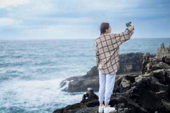 A caucasian person standing on rugged coastal rocks, capturing a selfie with the expansive ocean view in the background. Portugal
