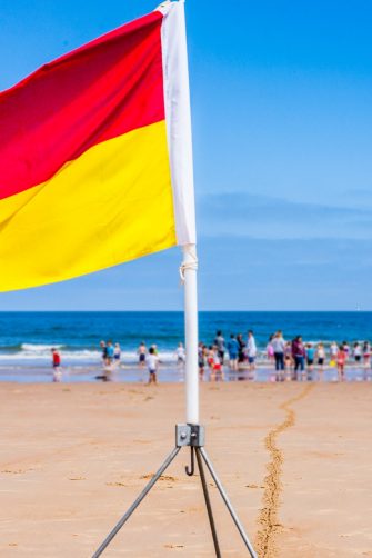 A red and yellow lifeguard safety flag on a beach with families and children in the background playing in the sea