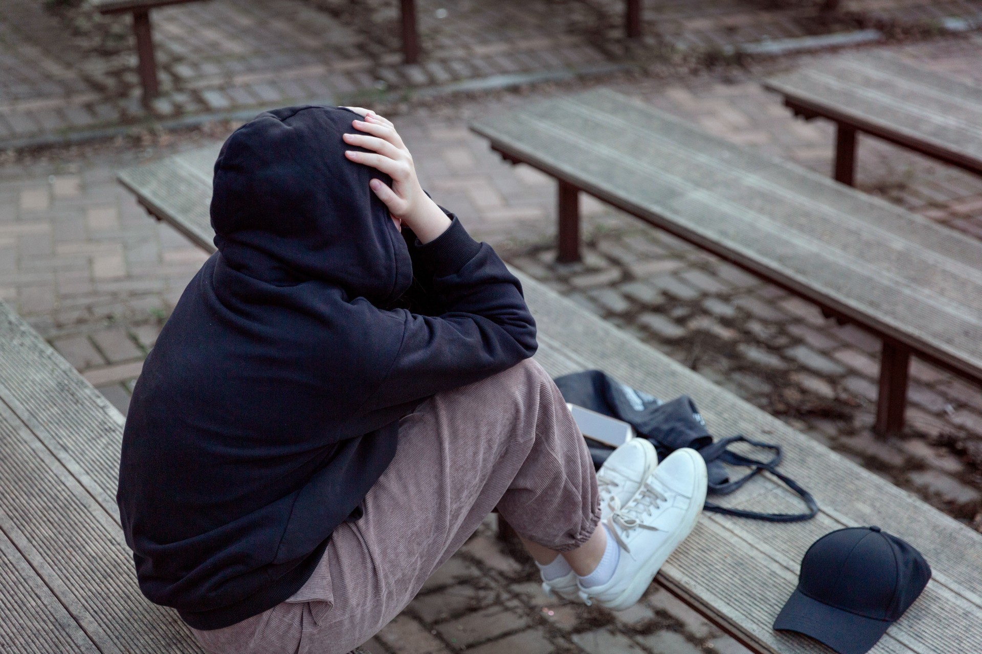 Teenager in a black hoodie with a hood on is sitting on a bench, covering face with her hands, back view