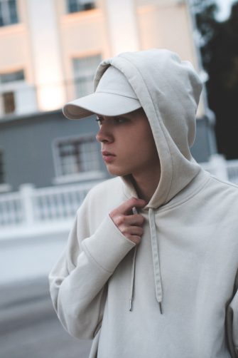 Young teenage boy 18-19 year old wear trendy beige hoodie and cap work out over city background early in morning in dark. Selective focus. Teenagerhood. Active lifestyle. Sad teenager have bad mood. 